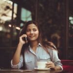 Chica asiática sonriente sentada en la mesa de una cafetería se toma un café mientras habla por teléfono