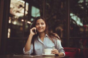 Chica asiática sonriente sentada en la mesa de una cafetería se toma un café mientras habla por teléfono