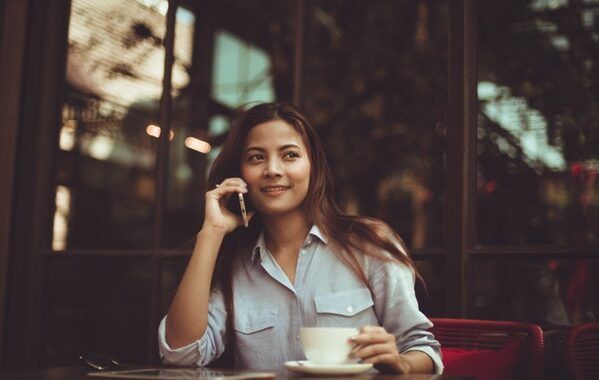 Chica asiática sonriente sentada en la mesa de una cafetería se toma un café mientras habla por teléfono