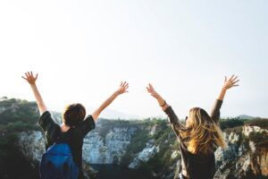 Dos chicas de espaldas alzan las manos en señal de victoria mientras llevan unas mochilas en un entorno natural rocoso