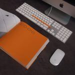 libreta naranja abierta junto a ordenador mac, sobre la mesa de un escritorio