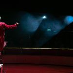 Vista de espaldas de un actor vestido de rojo que saluda hacia la oscuridad del patio de butacas