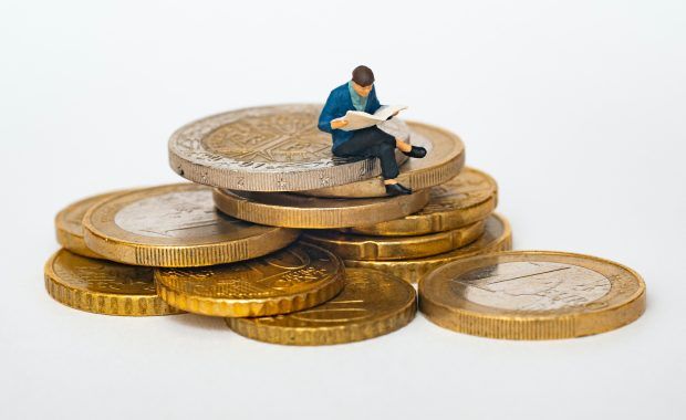 Una muñeco en miniatura de un señor leyendo un periódico está sentada sobre un pequeño puñado de monedas de euro y céntimos