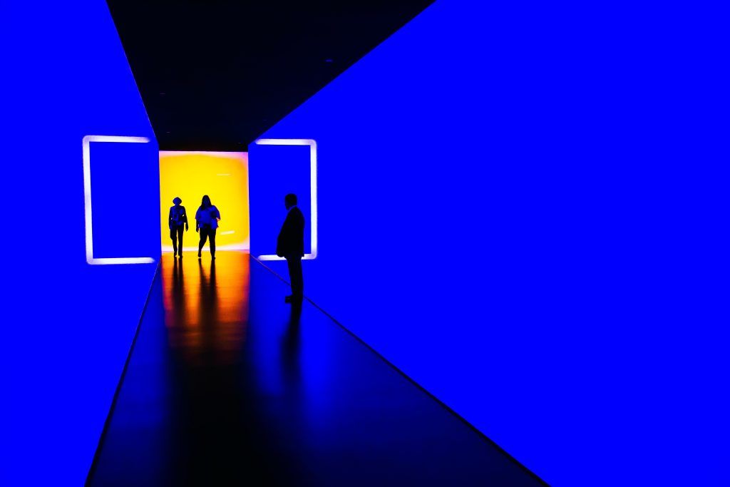 Una pasillo minimalista oscuro iluminado por intensa luz azul; el fondo del pasillo está iluminado en amarillo, y hay tres personas allí