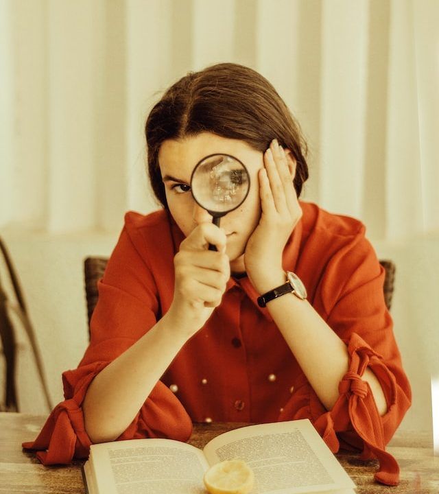 Mujer posa mirando a través de una lupa sentada frente a un libro