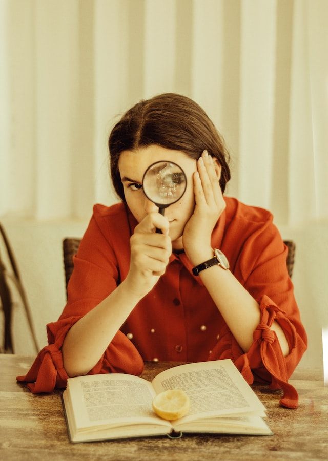 Mujer posa mirando a través de una lupa sentada frente a un libro